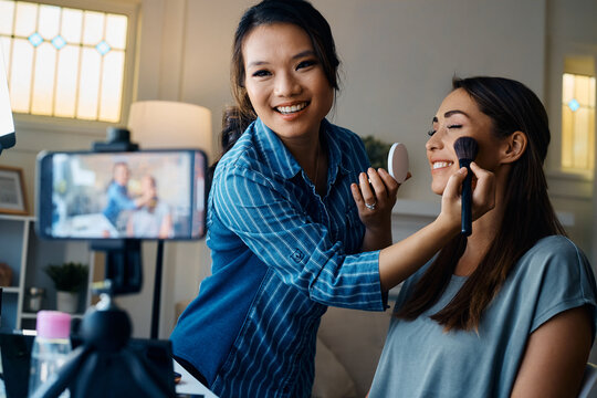 Happy  Asian beautician recording video while applying make up on young woman's face.