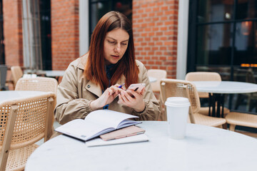 Young woman enjoying a coffee, sitting on the cafe terrace on the modern city street. Female worker using on mobile Phone standing outdoors. Person sitting at table and taking notes in notebook