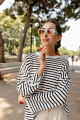 Cheerful happy elegant woman with dark hair wearing bright glasses and striped shirt is touching her chin and looking straight