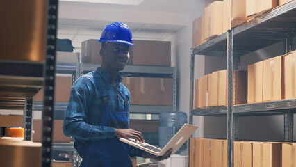 African american worker counting products on shelves, checking stock list on laptop before shipping order. Male person with hardhat using pc to work on merchandise inventory. Handheld shot.