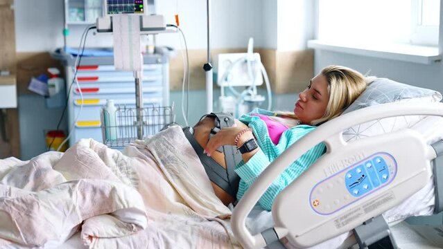 Blonde pregnant woman strokes her belly with attached cardiotocography belt. Future mom before childbirth in maternity hospital.
