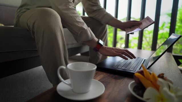 Closeup footage of male person holding passport in one hand and typing on laptop by another one sitting on the balcony. Caucasian man inputs his personal information into internet. Authorization theme
