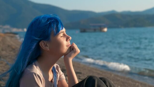 Fashioned girl on restless sea. A view of dreaming teen girl with blue hair relax on the shore in loneliness. A concept of luxury rest by the blue sea.