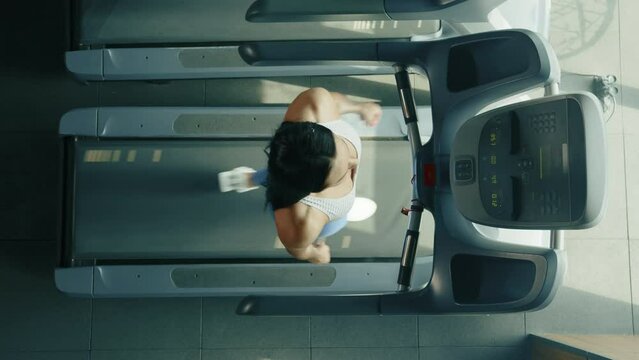 Young athletic brown-haired woman wearing white top, blue leggings, white sneakers walks on treadmill in gym. Top view. Monitor indicates results. High quality 4k footage