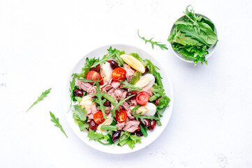 Nicoise salad with tuna, red tomatoes, boiled eggs, green beans and olives on plate, white table background, top view
