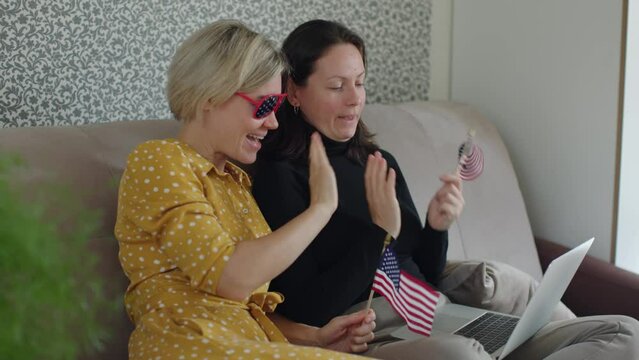 two female couple celebrating 4th july independence day online video call family laptop happy talking looking screen waving usa flags hands. stars and stripes on glasses have fun indoors home sit safa