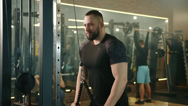 Slow motion medium shot of athletic young man focused on training on vertical row machine in gym. Man and woman exercising with barbells in the background. High quality 4k footage