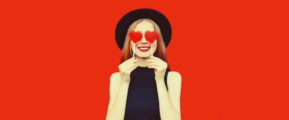 Portrait of beautiful young woman covering her eyes with red heart shaped lollipop wearing black round hat on background