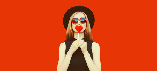 Portrait of beautiful young woman with lollipop blowing her lips with lipstick sending sweet air kiss wearing red heart shaped sunglasses, black round hat on background