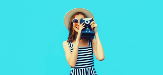Summer portrait of happy smiling young woman photographer with film camera wearing straw hat, dress, sunglasses on blue background