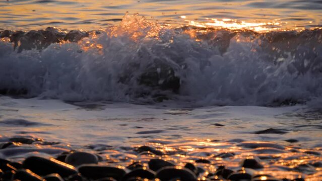 Slow motion, front view - sea water waves are crashing on the pebble beach at sunset - close up. Nature and relaxation concept