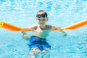 Oudoor summer activity. Concept of fun, health and vacation. Happy smiling boy eight years old in swim glasses swim in the pool with noodle in hot summer day.