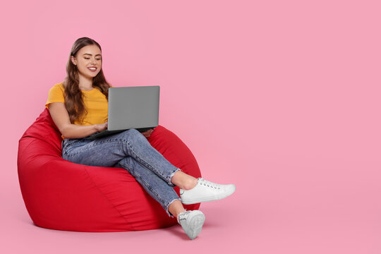 Happy woman with laptop sitting on beanbag chair against pink background. Space for text