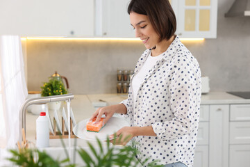 Happy young woman washing plate above sink in modern kitchen