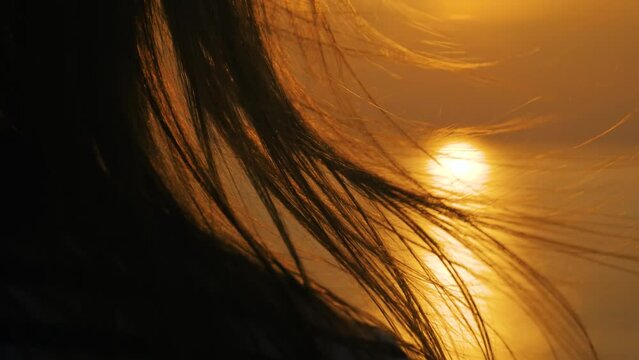 Slow motion: hair of woman is fluttering in the wind against warm sunset sky with sun lens flares, close up. Abstract, freedom and summer concept