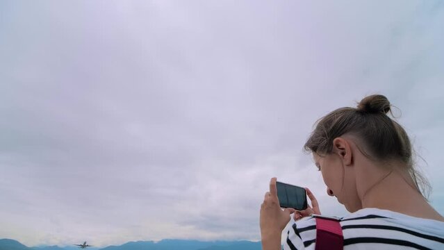 Slow motion: woman holds smartphone, takes photo or shoots video of departing passenger airplane, airliner against the gray sky - wide angle view. Photography, plane spotting and technology concept