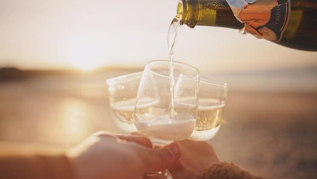 Sparkling wine in glasses on the ocean beach at sunset. Pouring alcoholic drink from the bottle into clear glasses, picnic on the seaside in summer. Party outdoors, happy moments, lens flare.