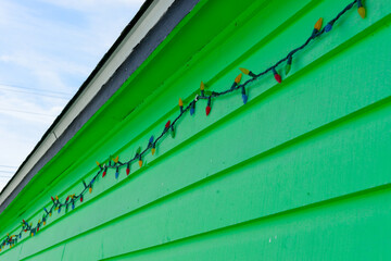 Obraz premium The exterior vibrant green wooden clapboard on the wall of a storage shed. A string of multi-colored Christmas lights is strung just below a flat shingled roof. 