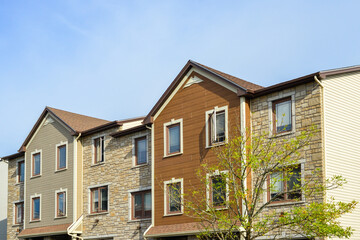 The top floors of a beige colored townhouse with vinyl siding, brown stone veneer, glass windows, asphalt shingles, and peaked roofs. There are tall trees with leaves in front of the modern building. 