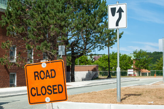 An orange colored road sign with road closed in black lettering. The sign is in the foreground with a road construction site in the background and a large brick building with tall green trees.