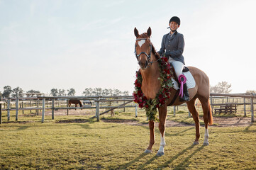 A couple of winners. Full length shot of a young female jockey sitting on her wreath-wearing horse...