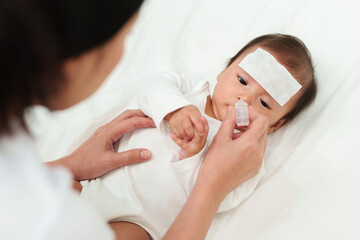 sick baby with cool fever pad gets nose drops by mother on bed