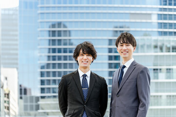オフィスビルの建物の前で微笑むカメラ目線の20代と30代のスーツを着たビジネスマンの男性2人