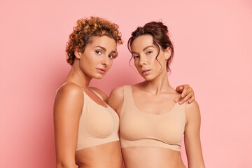 Portrait shot of two girlfriends inside the studio, curly one resting her hand on the shoulder of her friend, both dressed in beige sport bras, friendship concept, copy space, high quality photo - obrazy, fototapety, plakaty