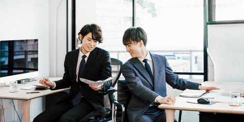 オフィスで同僚と背中合わせで振り向いて相談しながら仕事の打ち合わせをして働く男性2人