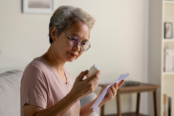 Asian senior woman sitting on sofa in her living room reading the information sheet of her prescribed medicine - 596529288