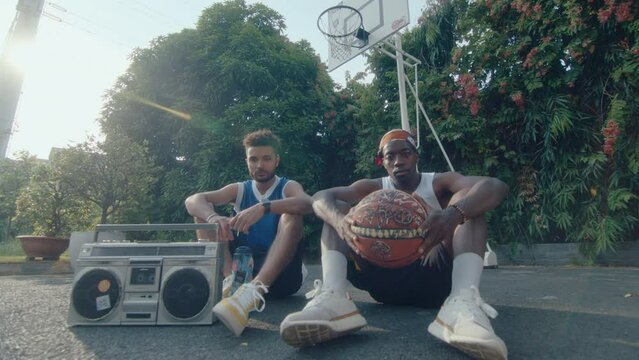 Two young male friends in sportswear sitting with basketball and ghetto blaster on outdoor playground, posing on camera with confidence. Full shot, handheld camera