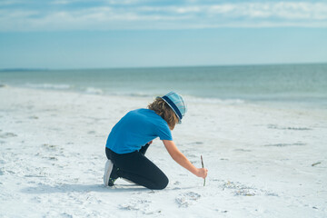Summer vacation concept. Happy little kid drawing a on sand on sandy beach. Travel and adventure kids concept. Lovely kid play outdoor. Pretty little child in hat relaxing on the beach drawing in the
