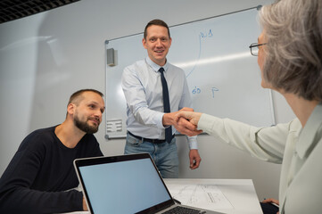 The boss makes a presentation to subordinates at the white board. Caucasian man shaking hands with middle aged woman. 