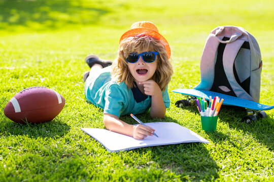 Creativity children concept. Clever school boy doing homework, writing on copy book in green grass of park. Back to school, knowledge, education, learning concepts.