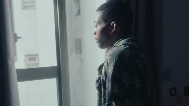 Young black man putting on casual shirt and looking through the window, getting dressed before leaving home. Medium shot