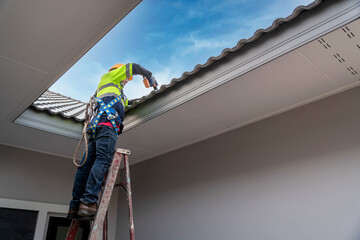 Asian roofer wearing safety harness belt using air or pneumatic nail gun and installing house roof...