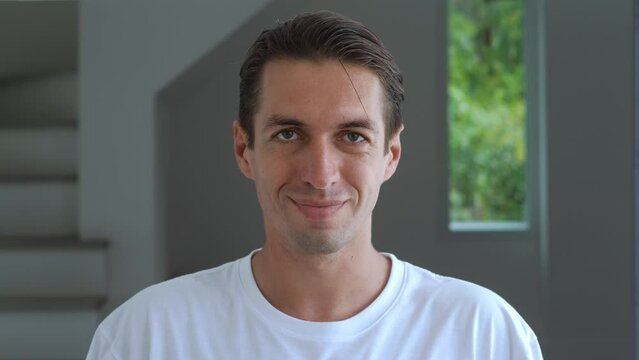 Close-up Portrait Of A Millennial Man In A White T-shirt Looks At The Camera And Smiles Against The Backdrop Of A Home Interior Or Office. Satisfied Man 30 Years Old Looking At The Camera With A Smile