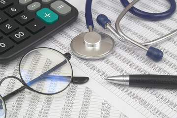 Documents with numbers, calculator, eyeglasses and stethoscope with pen close up.