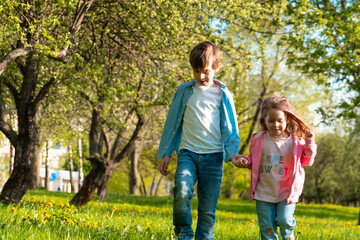 Adorable little girl with brother walking together among trees in green park while holding hands