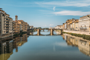 Fototapeta na wymiar Gorgeous view of an old stone bridge across a river in Italy on a sunny warm summer evening on a blue sky. The concept of historical important infrastructural structures in Europe. Copyspace