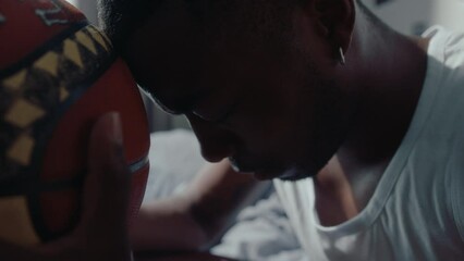Young black basketball player sitting in his bedroom, touching ball with forehead, getting nervous before a game and trying to focus. Close-up shot, selective focus