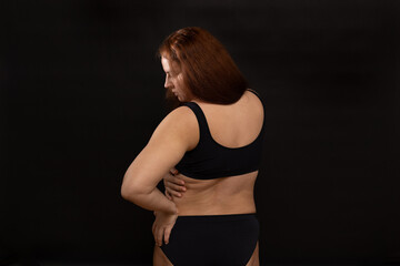 Fototapeta na wymiar Plus size woman in black underwear turning to side, back view. Body fat folds. Flaunt figure imperfections. Studio portrait over black background. Concept of obesity, body positive, self acceptance.