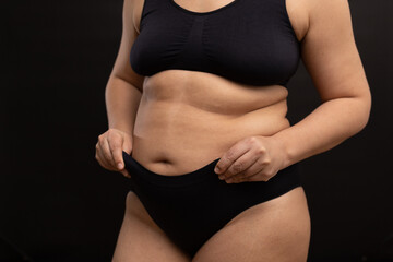 Overweight woman in black underwear pull up panties on belly. Flaunt figure imperfections, fat folds on body. Studio portrait over black background. Concept of obesity, body positive, self acceptance.