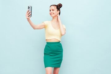 Naklejka premium Young beautiful smiling female in trendy summer yellow t-shirt and green skirt. Carefree woman with two horns hairstyle posing near blue wall in studio. Positive model having fun. Taking selfie photos