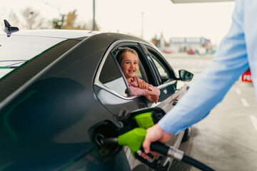 The daughter sits in the car leaning against the window and watches her father pour fuel into the...