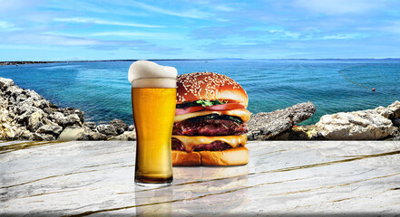 Glass of beer and a hamburger on white and gold marble with mediterranean seascape in the background