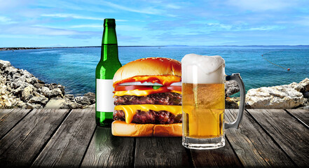 Mug and beer bottle and a hamburger on white and gold marble with mediterranean seascape in the background