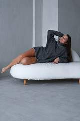 Brunette woman in warm sweater with bare legs relaxing. Stylish woman with long hair sitiing on white sofa. Vertical