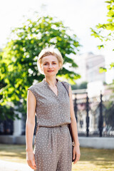 Portrait of wonderful looking woman in elegant costume standing outdoors among green trees in alley with cute smile. Middle aged lady photo on sunny day. Selective focus, blurred urban background.