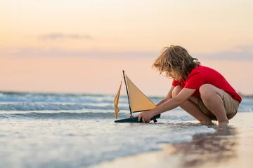 Fotobehang Kid boy playing with toy boat in sea water. Happy holiday by the sea. Kid dreaming about sailing. Happy kid playing with toy sailing boat. Summer vacation. Cute little kid hold toy sailboat. © Volodymyr